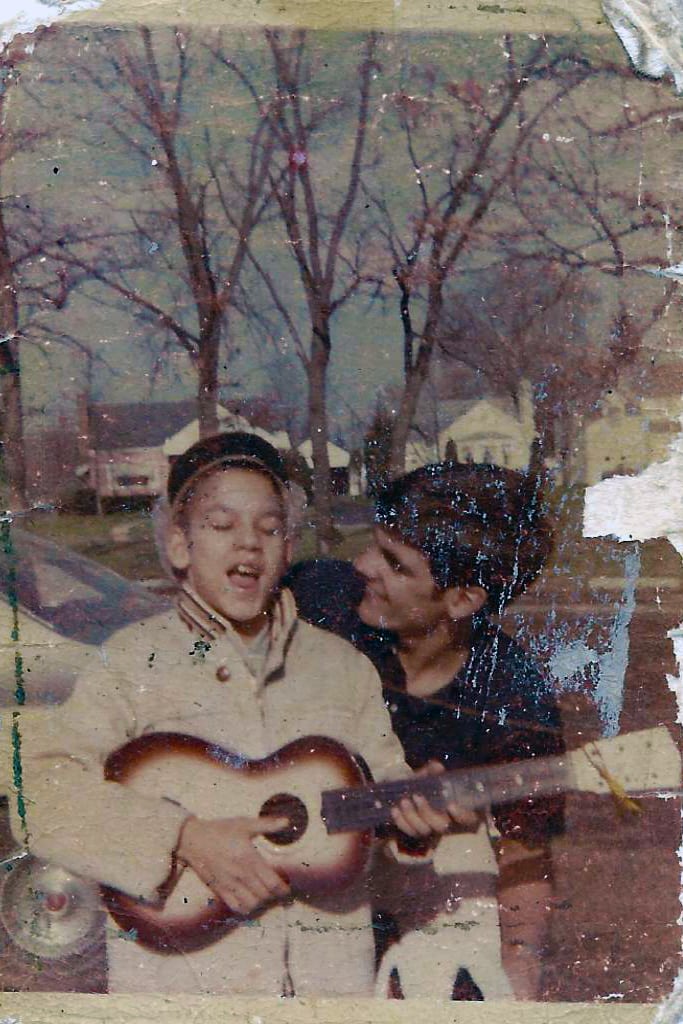 A creased family photograph of Jeff Moyer as a boy, singing as he plays a small guitar, his brother Mark smiling at his side.