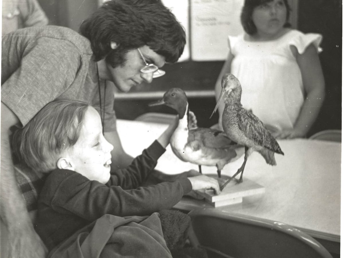 A black-and-white photograph from 1974: a young Jeff Moyer leans in beside a smiling child who reaches out to touch taxidermied birds on a classroom table.