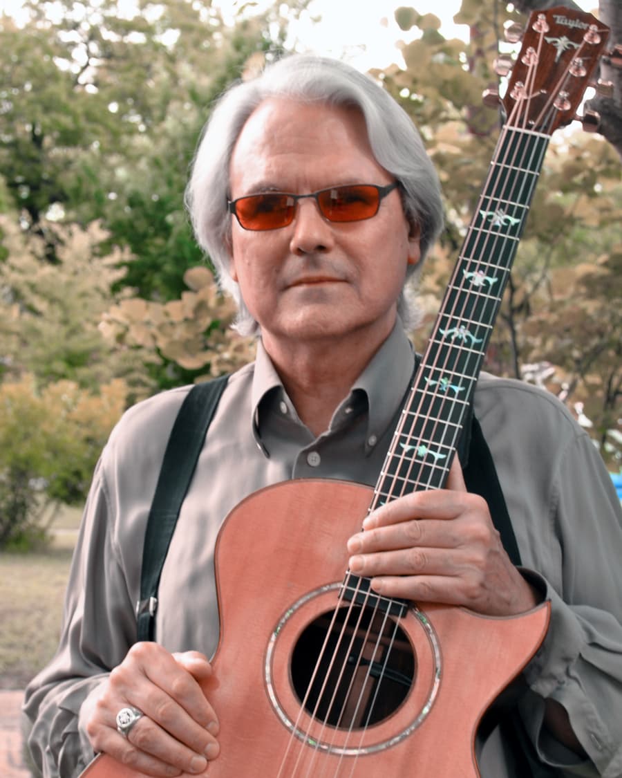 Jeff Moyer, silver-haired in tinted glasses, stands among trees holding his Taylor guitar upright against his chest.