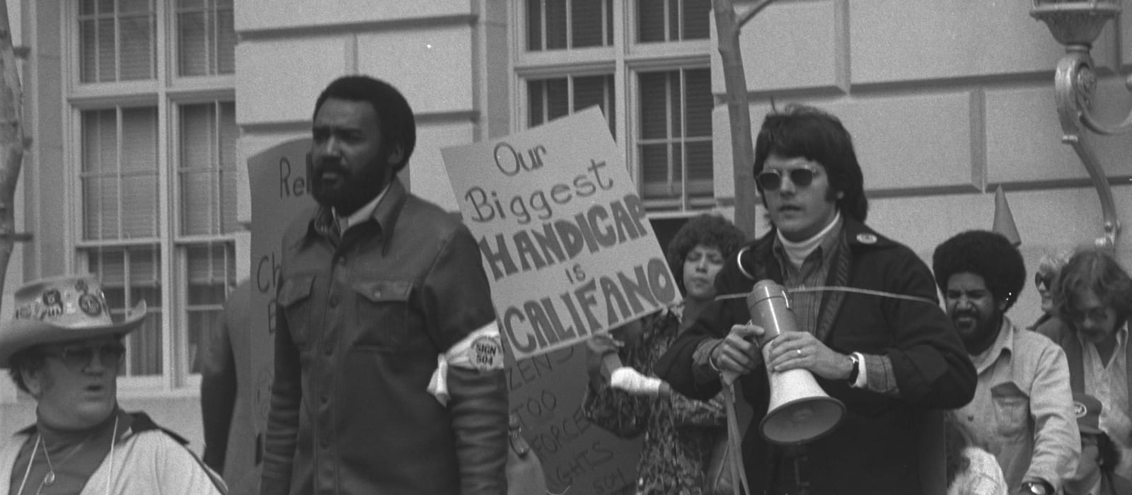 A black-and-white photograph of demonstrators outside a government building during the 1977 Section 504 protests. A young man in dark glasses holds a megaphone beside a hand-lettered sign reading 'Our biggest handicap is Califano.'
